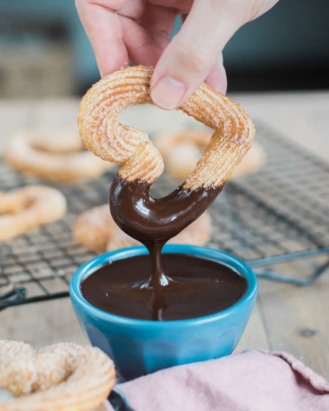 Honeywell Bakes Churros Kit For Baking - Person dipping a churro into a bowl of chocolate sauce on a cooling rack with more churros in the background.