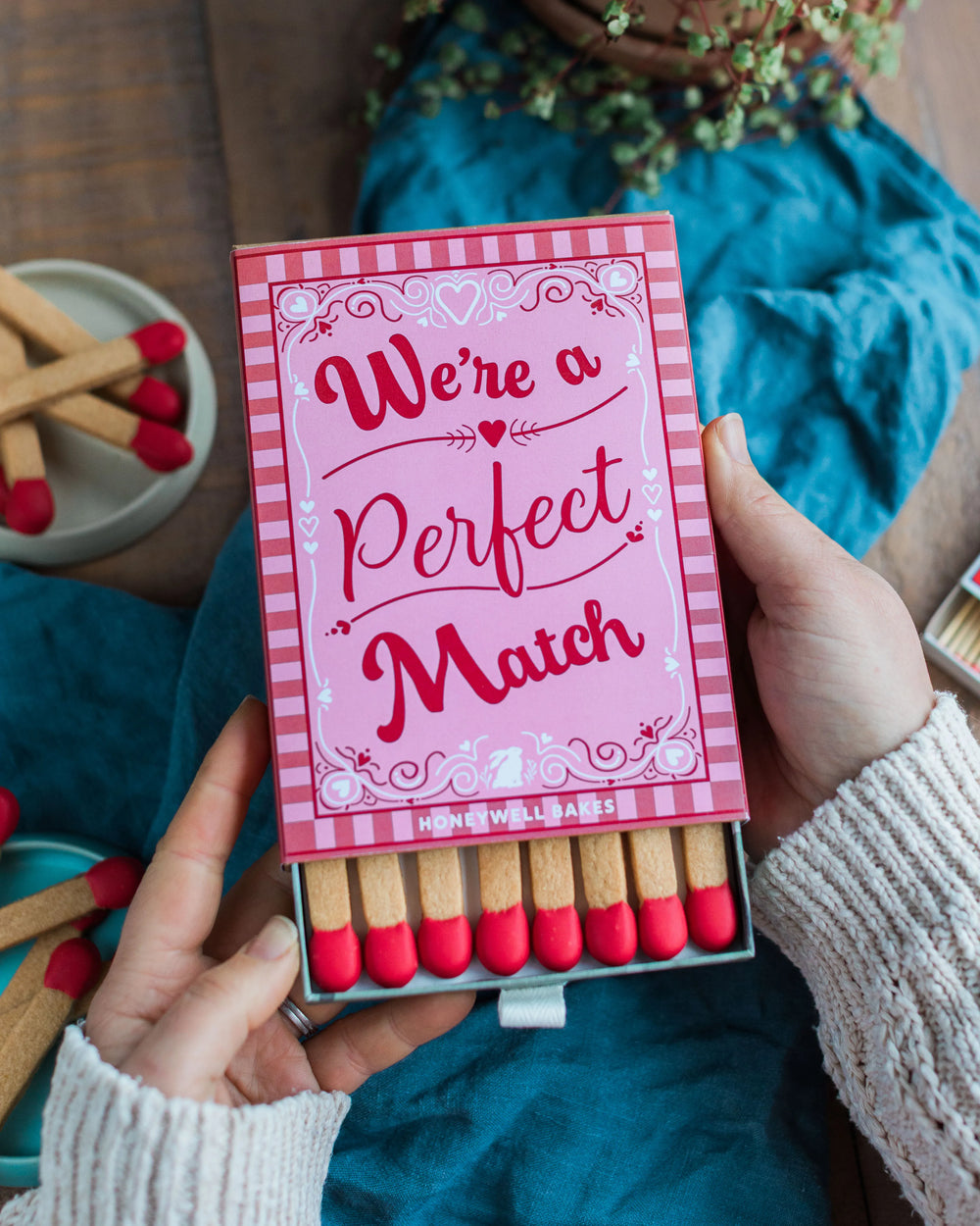 Honeywell Bakes A Perfect Match - A Perfect Match Biscuit Gift - Shortbread Cookie Matches with a pink box labeled 'We're a Perfect Match' on a wooden surface with blue fabric.