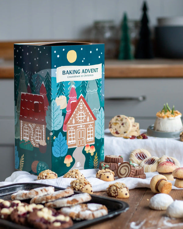 Honeywell Bakes Bakery Advent Calendar - 12 Days Of Cosy Baking - A festive 'Baking Advent' gift box for Christmas is displayed on a wooden table surrounded by an assortment of baked cookies, pretzels, and a small cake. The packaging features a dark teal winter forest illustration with gingerbread houses and a snow moon.