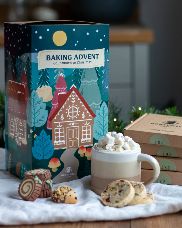 Honeywell Bakes Bakery Advent Calendar - 12 Days Of Cosy Baking - A festive Baking Advent gift set box by Honeywell Bakes styled on a white cloth with a mug of hot cocoa topped with marshmallows, cookies, and assorted chocolates in the foreground.