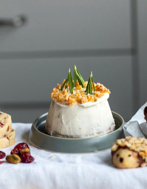 Honeywell Bakes Bakery Advent Calendar - 12 Days Of Cosy Baking - A small frosted dessert or cake on a gray plate, topped with crumbled nuts and green sprigs, set on a white cloth with cookies and dried cranberries in the foreground.