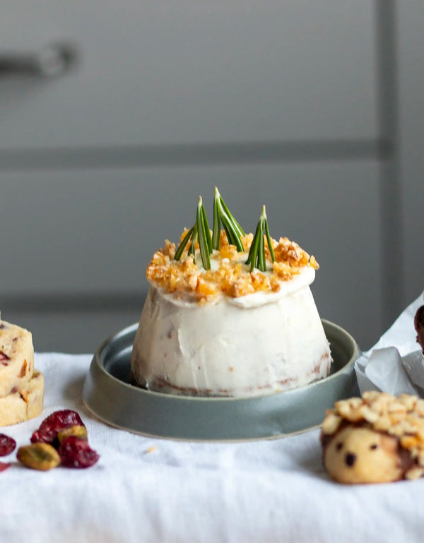 Honeywell Bakes Bakery Advent Calendar - 12 Days Of Cosy Baking - A small frosted dessert or cake on a gray plate, topped with crumbled nuts and green sprigs, set on a white cloth with cookies and dried cranberries in the foreground.