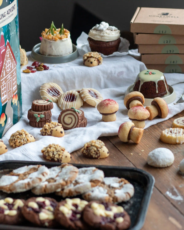 Honeywell Bakes Bakery Advent Calendar - 12 Days Of Cosy Baking - An assortment of baked goods displayed on a rustic wooden table, including decorated cakes, cookies, and pastries, with a stack of brown boxes and a colourful printed box visible in the background.