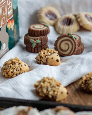Honeywell Bakes Bakery Advent Calendar - 12 Days Of Cosy Baking - Decorated Christmas cookies arranged on a white cloth and baking tray, including log cake cookies, hedgehog cookies with chopped nuts, and striped sugar cookies. A branded box is partially visible on the left.