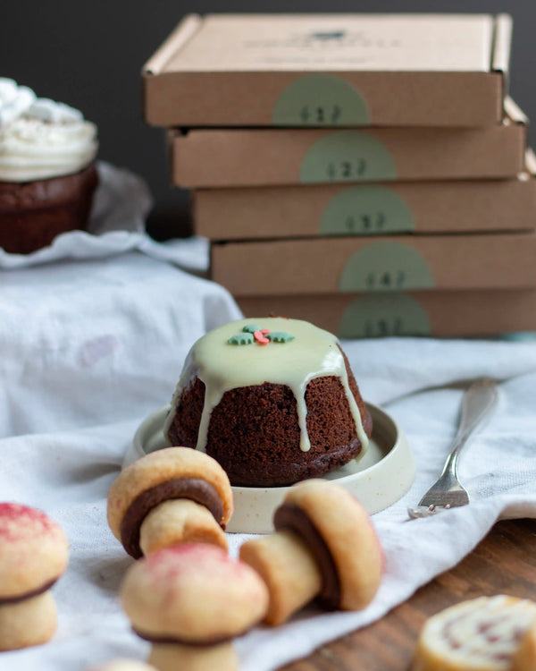 Honeywell Bakes Bakery Advent Calendar - 12 Days Of Cosy Baking - A small chocolate cake with pale green icing sits on a plate on a tablecloth, surrounded by cookies, with stacked cardboard boxes in the background.