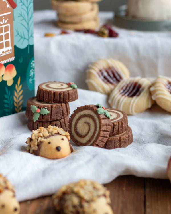 Honeywell Bakes Bakery Advent Calendar - 12 Days Of Cosy Baking - A styled flat-lay of assorted cookies on a white cloth, including log-shaped swirled cookies, a hedgehog-shaped cookie, and jam-filled cookies, with a decorative green box partially visible in the background.