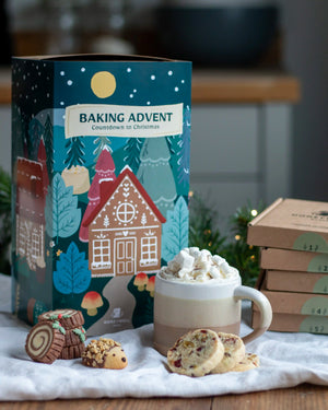Honeywell Bakes Bakery Advent Calendar - 12 Days Of Cosy Baking - A festive Baking Advent box for Christmas displayed on a white cloth with baked goods and a mug of hot cocoa. The illustrated box features gingerbread houses and trees under a starry night sky.