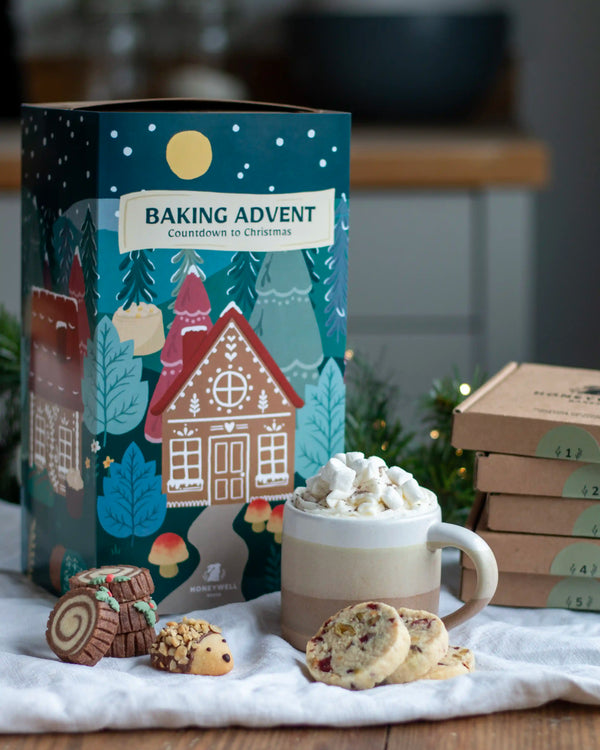 Honeywell Bakes Bakery Advent Calendar - 12 Days Of Cosy Baking - A festive Baking Advent box for Christmas displayed on a white cloth with baked goods and a mug of hot cocoa. The illustrated box features gingerbread houses and trees under a starry night sky.
