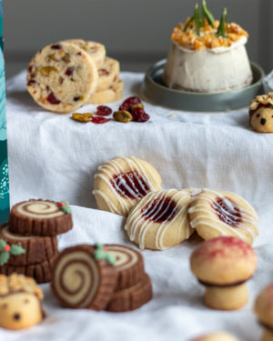 Honeywell Bakes Bakery Advent Calendar - 12 Days Of Cosy Baking - An assortment of decorated cookies arranged on a white fabric surface, including swirled log cookies, jam cookies with white icing, and small festive cookies, with a small dessert in the background.