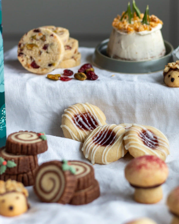 Honeywell Bakes Bakery Advent Calendar - 12 Days Of Cosy Baking - An assortment of decorated cookies arranged on a white fabric surface, including swirled log cookies, jam cookies with white icing, and small festive cookies, with a small dessert in the background.