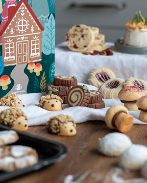 Honeywell Bakes Bakery Advent Calendar - 12 Days Of Cosy Baking - A styled food product display featuring a colourful Honeywell gingerbread house box alongside assorted baked cookies, rolls, and pastries arranged on a wooden table with a white cloth.