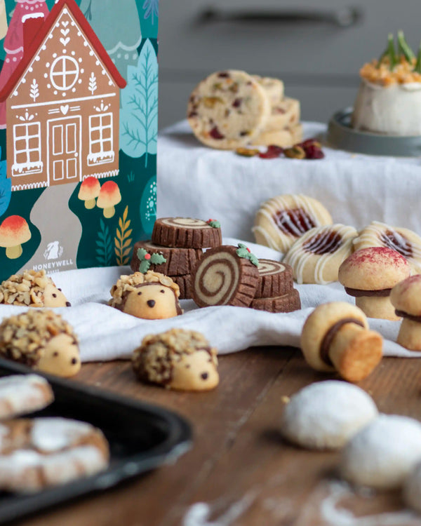 Honeywell Bakes Bakery Advent Calendar - 12 Days Of Cosy Baking - A styled food product display featuring a colourful Honeywell gingerbread house box alongside assorted baked cookies, rolls, and pastries arranged on a wooden table with a white cloth.