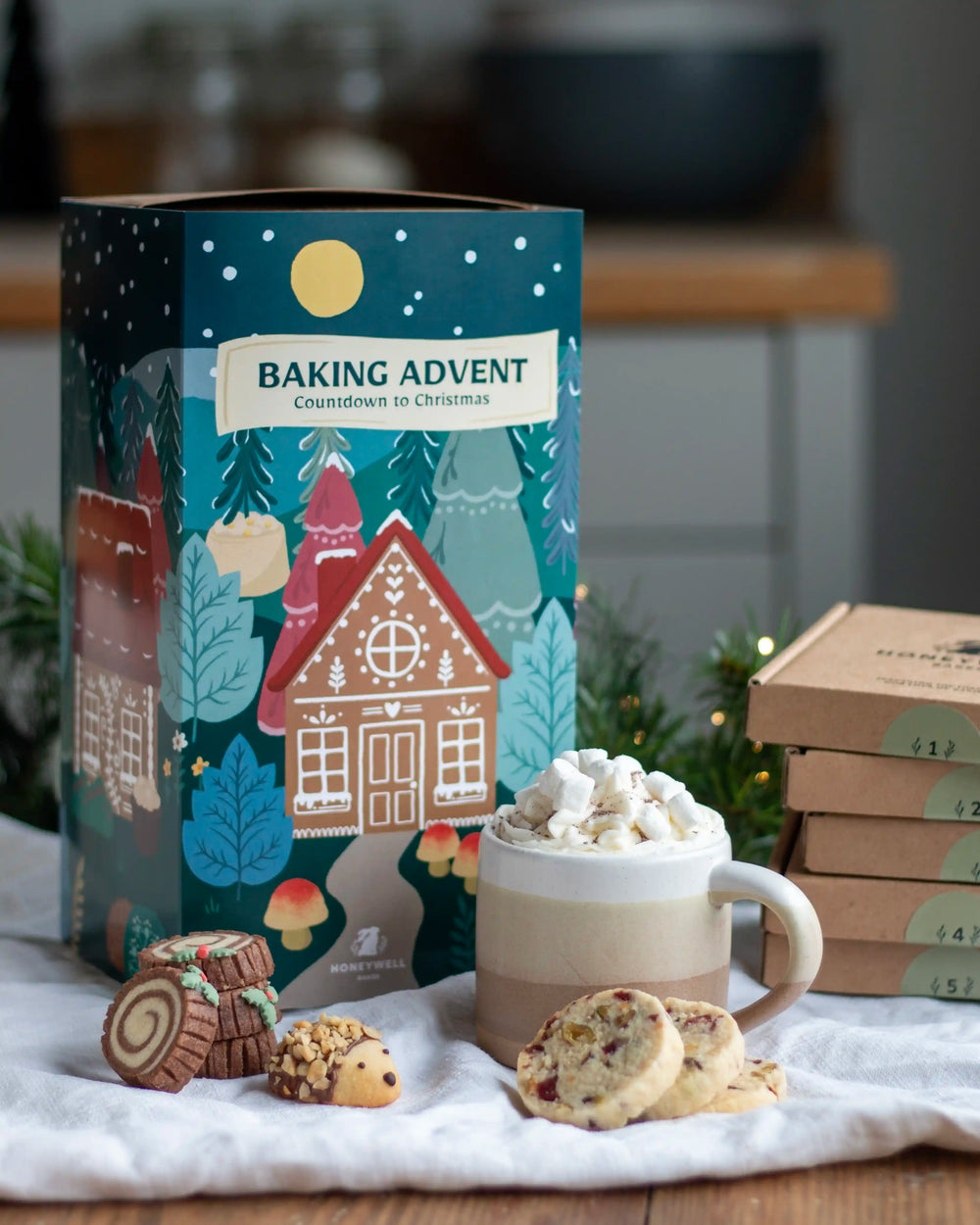 Honeywell Bakes Bakery Advent Calendar - 12 Days Of Cosy Baking - A festive baking advent box labeled 'BAKING ADVENT Countdown to Christmas' stands on a table with cookies, a mug of hot chocolate topped with marshmallows, and several smaller boxes in the background. The scene is styled in a warm kitchen-like setting.