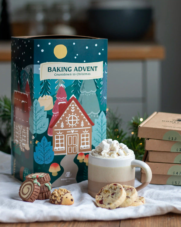 Honeywell Bakes Bakery Advent Calendar - 12 Days Of Cosy Baking - A festive baking advent box labeled 'BAKING ADVENT Countdown to Christmas' stands on a table with cookies, a mug of hot chocolate topped with marshmallows, and several smaller boxes in the background. The scene is styled in a warm kitchen-like setting.