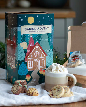 Honeywell Bakes Bakery Advent Calendar - 12 Days Of Cosy Baking - A festive Baking Advent countdown box with Christmas tree and gingerbread house artwork, styled with a mug of whipped-cookie-drink, cookies, and an open box containing Nutella and a biscuit on a white cloth.