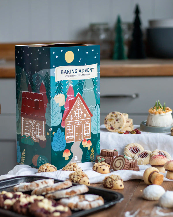 Honeywell Bakes Bakery Advent Calendar - 12 Days Of Cosy Baking - A festive 'Baking Advent' gift box decorated with a Christmas village illustration, set on a wooden table surrounded by assorted baked cookies and pastries in a softly blurred kitchen background.