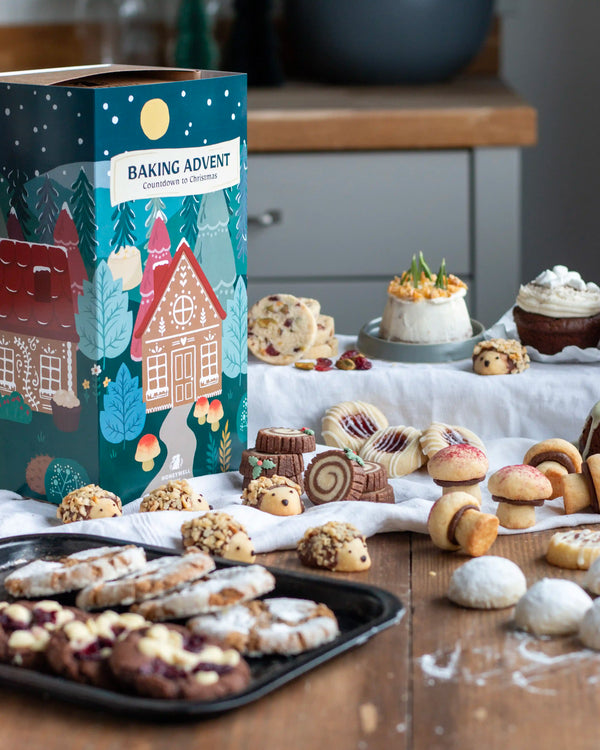 Honeywell Bakes Bakery Advent Calendar - 12 Days Of Cosy Baking - A decorative baking advent box labeled 'BAKING ADVENT' stands on a wooden table beside an assortment of cookies, pastries, and a small cake in a kitchen setting.