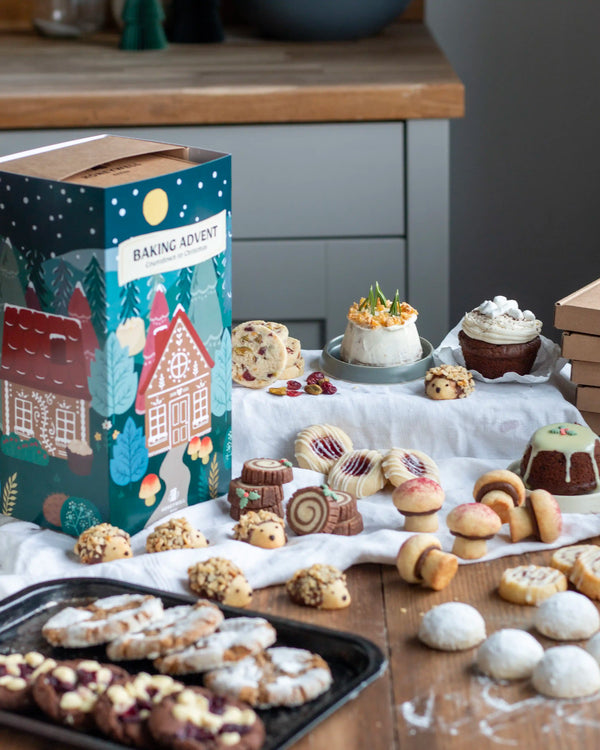 Honeywell Bakes Bakery Advent Calendar - 12 Days Of Cosy Baking - A festive Baking Advent box with a winter village illustration sits on a wooden table surrounded by assorted baked cookies, cupcakes, and a small cake. The scene is styled in a kitchen setting with a wooden countertop and gray cabinets in the background.