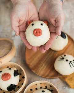 Honeywell Bakes Bao Buddies Baking Kit - Hands holding a small bao bun shaped like a pig with other similar bao on a wooden board.