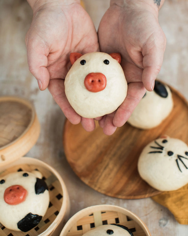 Honeywell Bakes Bao Buddies Baking Kit - Hands holding a small bao bun shaped like a pig with other similar bao on a wooden board.