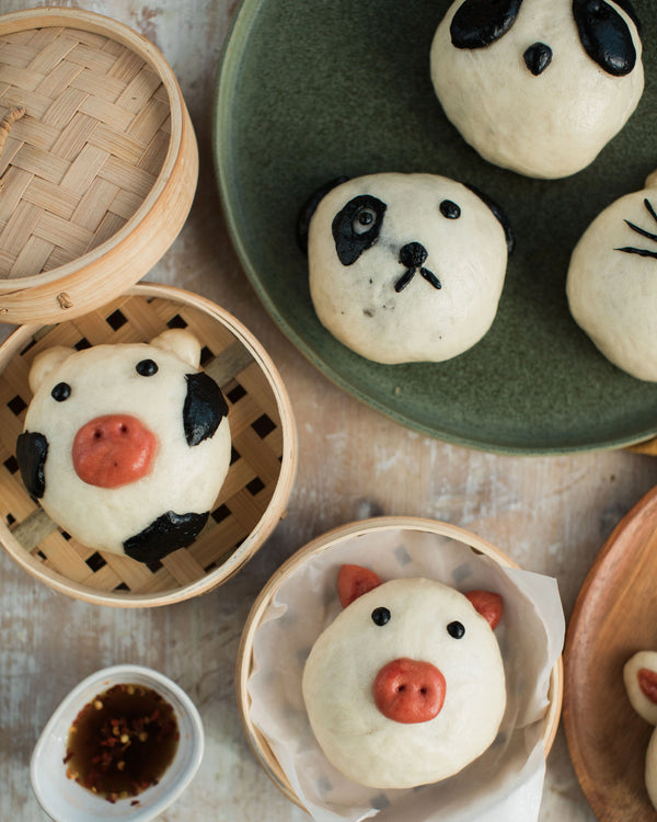 Honeywell Bakes Bao Buddies Baking Kit - Panda-shaped buns on a wooden table with a bamboo steamer and small bowl of dipping sauce.