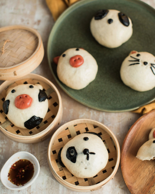 Honeywell Bakes Bao Buddies Baking Kit -  Animal-shaped bao buns on a plate with a wooden steamer and small bowl of sauce.