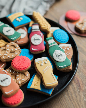  Honeywell Bakes Beer Lover’s Biscuit Jar - Assorted decorated cookies shaped like bottles and other items on a black plate.