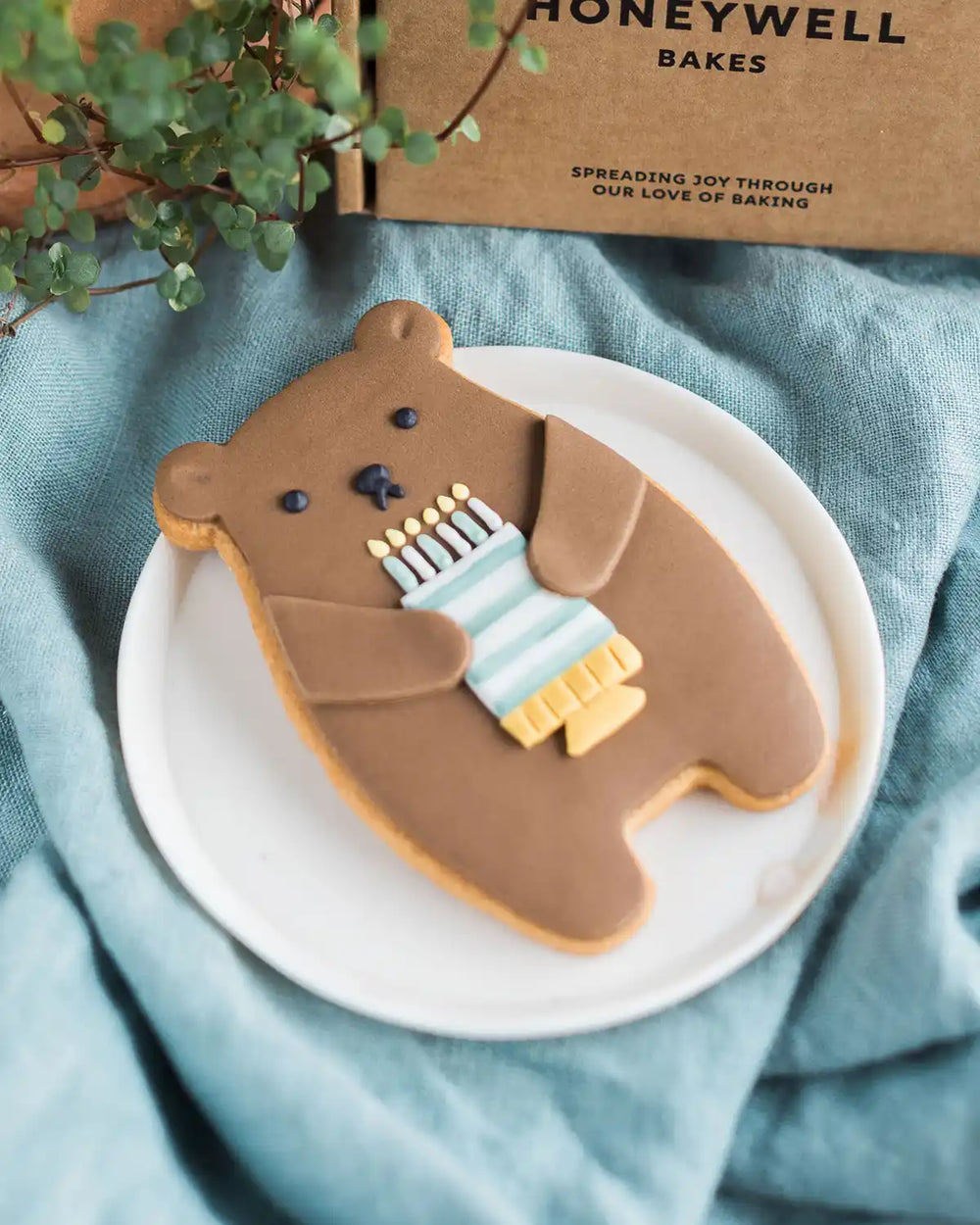 Honeywell Bakes Birthday Bear Biscuit Bear-shaped cookie holding a birthday cake, placed on a blue cloth with a 'Honeywell Bakes' box in the background.
