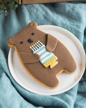 Honeywell Bakes Birthday Bear Biscuit Bear - Brown bear-shaped cookie holding a birthday cake on a white plate with a blue cloth background