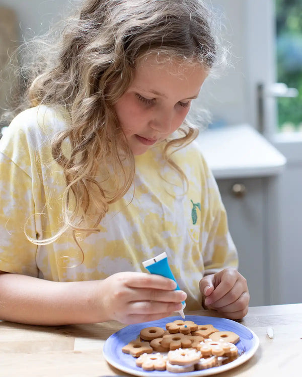 Honeywell Bakes Biscuit Jewellery Craft Kit - Child sitting at a table with a plate of cookies and a blue piping tube.