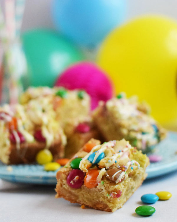 Honeywell Bakes Chocolate Rainbow Blondie Baking Kit  - colourful blondie bars with smarties on a blue plate against a festive background with balloons and straws.