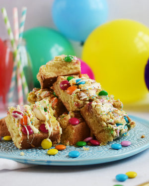 Honeywell Bakes Chocolate Rainbow Blondie Baking Kit  - colourful blondie bars with smarties on a blue plate against a festive background with balloons and straws.