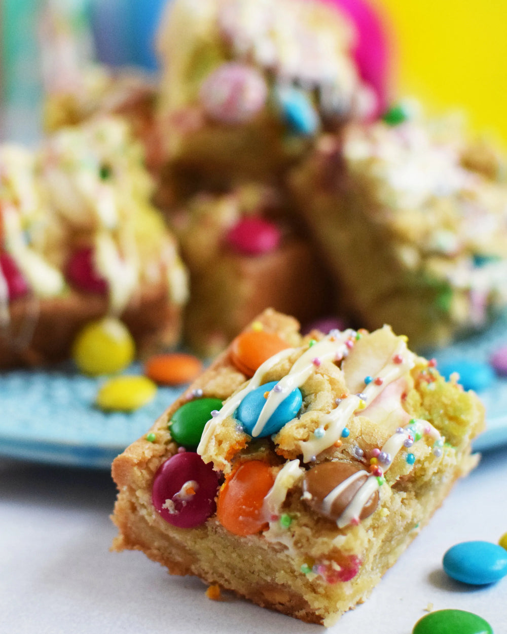 Honeywell Bakes Chocolate Rainbow Blondie Baking Kit  - colourful blondie bars with smarties on a blue plate against a festive background with balloons and straws.
