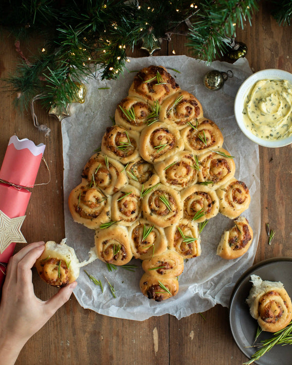 Honeywell Bakes - A hand is holding a cheese and tomato roll shaped like a Christmas tree, served with a side of holiday dip on a rustic wooden table decorated with pine branches and lights.