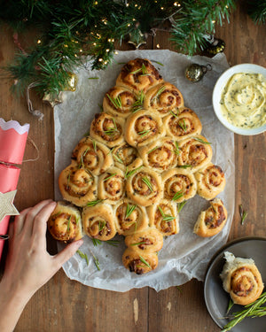 Honeywell Bakes - A tree-shaped bread arrangement made of rolled dough pieces with rosemary sprigs, served on parchment paper on a wooden table with holiday decorations, a dipping sauce bowl, and a hand reaching in.