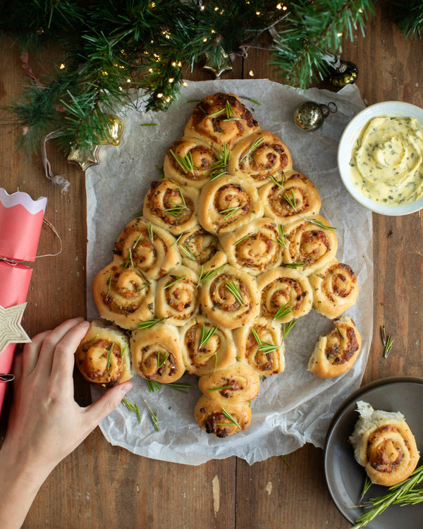 Honeywell Bakes - A tree-shaped bread arrangement made of rolled dough pieces with rosemary sprigs, served on parchment paper on a wooden table with holiday decorations, a dipping sauce bowl, and a hand reaching in.