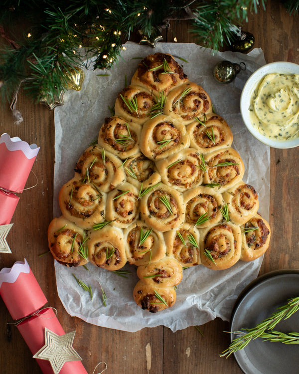 Honeywell Bakes - A Christmas tree-shaped bread made of rolled dough swirls, topped with rosemary, presented on parchment paper on a wooden table with holiday decorations, a bowl of dipping sauce, and crackers nearby.