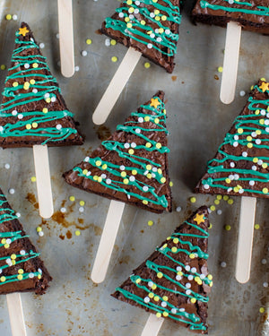 Honeywell Bakes Christmas Tree Brownie Baking Kit, Triangle-shaped brownie pops decorated to look like Christmas trees, with teal icing, yellow and white sprinkles, and star toppers on wooden sticks, arranged on a rustic metal surface.