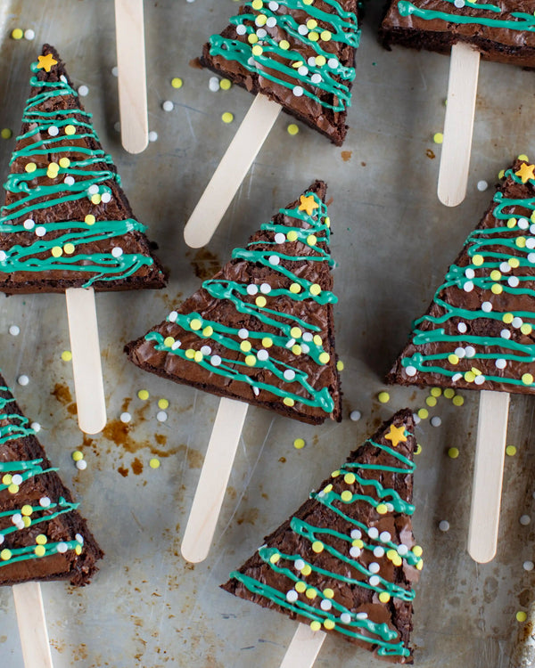 Honeywell Bakes Christmas Tree Brownie Baking Kit, Triangle-shaped brownie pops decorated to look like Christmas trees, with teal icing, yellow and white sprinkles, and star toppers on wooden sticks, arranged on a rustic metal surface.