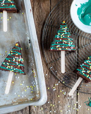 Honeywell Bakes Christmas Tree Brownie Baking Kit, Tree-shaped brownie pops decorated with teal icing and colourful sprinkles, arranged on a metal baking sheet and a wire cooling rack on a rustic wooden surface, with a small bowl of green icing in the corner.