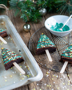 Honeywell Bakes Christmas Tree Brownie Baking Kit, Holiday-themed tree-shaped brownie pop baking kit, decorated with green icing and gold and white sprinkles, served on wooden sticks on a cooling rack and baking tray with festive pine branches and ornaments in the background.