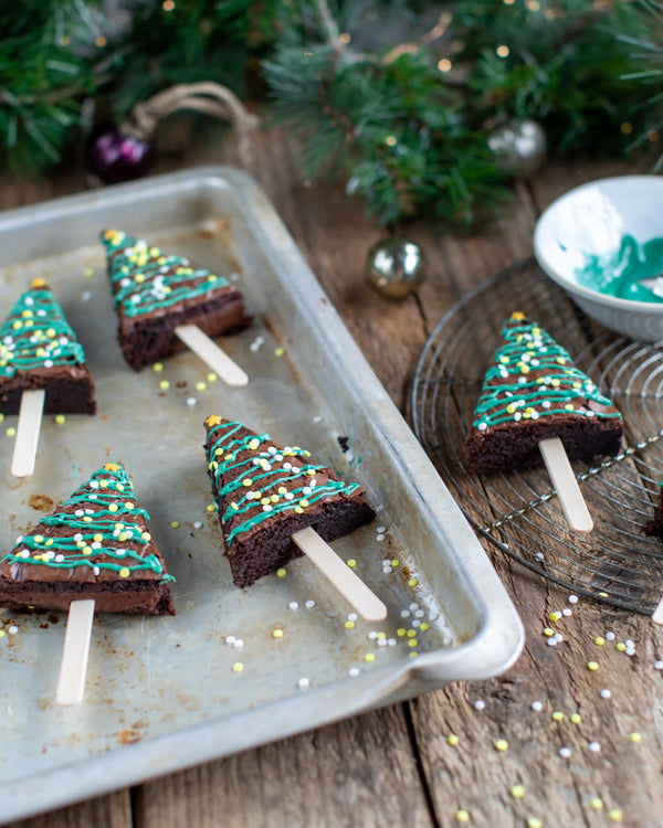 Honeywell Bakes Christmas Tree Brownie Baking Kit, Christmas tree-shaped brownie pops decorated with green frosting and gold and silver sprinkles, arranged on a baking tray and a cooling rack on a rustic wooden table with holiday greenery in the background.
