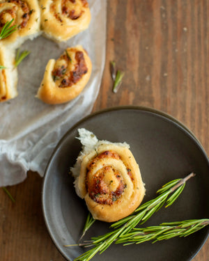 Honeywell Bakes - A baked cheese and tomato roll with herbs, served on a dark gray plate with a rosemary sprig on a rustic wooden table, with more rolls visible in the background on parchment paper.