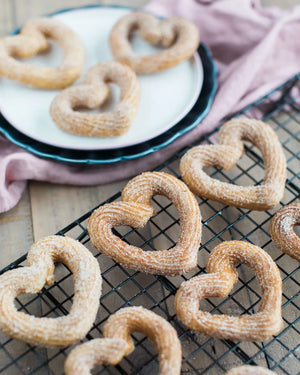Honeywell Bakes Churros Kit For Baking - Heart-shaped churros on a cooling rack with a plate of churros in the background.