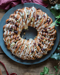 A Cinnamon Roll Wreath Baking Kit from Honeywell Bakes topped with white icing, dried cranberries, and chopped pistachios, served on a round blue plate over a rustic wooden table with holiday greenery and fabric.