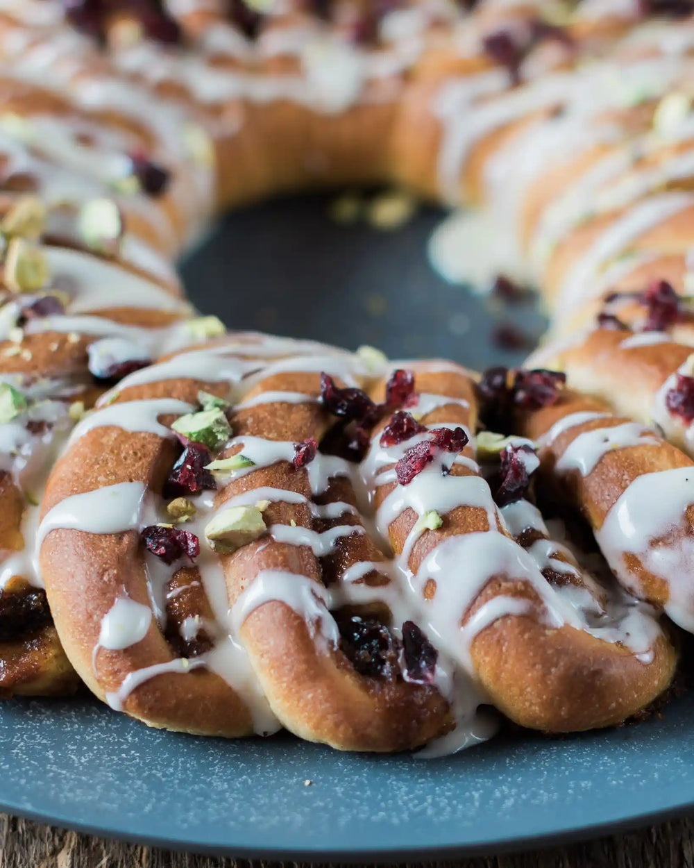 A Cinnamon Roll Wreath Baking Kit from Honeywell Bakes topped with white icing, dried cranberries, and chopped pistachios, served on a round blue plate over a rustic wooden table with holiday greenery and fabric.