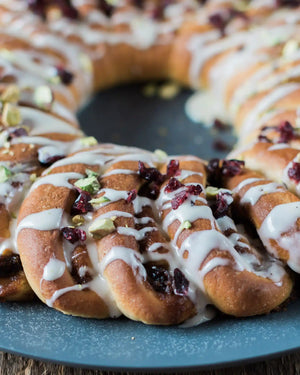 A Cinnamon Roll Wreath Baking Kit from Honeywell Bakes topped with white icing, dried cranberries, and chopped pistachios, served on a round blue plate over a rustic wooden table with holiday greenery and fabric.