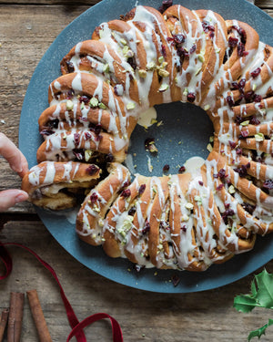 A Cinnamon Roll Wreath Baking Kit from Honeywell Bakes topped with white icing, dried cranberries, and chopped pistachios, served on a round blue plate over a rustic wooden table with holiday greenery and fabric.