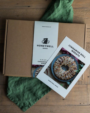 A Honeywell Bakes Cinnamon Roll Wreath bread baking kit shown with a kraft cardboard box, a printed recipe card, and a green cloth on a wooden tabletop.