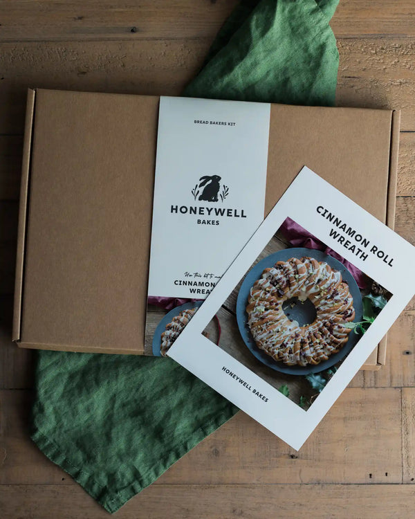A Honeywell Bakes Cinnamon Roll Wreath bread baking kit shown with a kraft cardboard box, a printed recipe card, and a green cloth on a wooden tabletop.